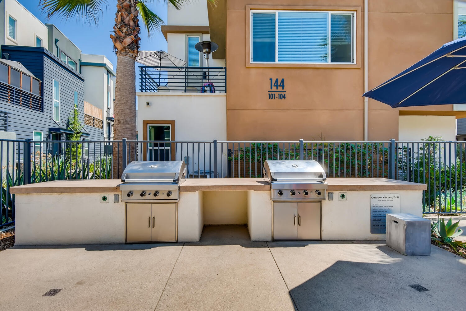An outdoor grilling area in a residential complex with stainless steel grills, a beige countertop, and a black metal fence.