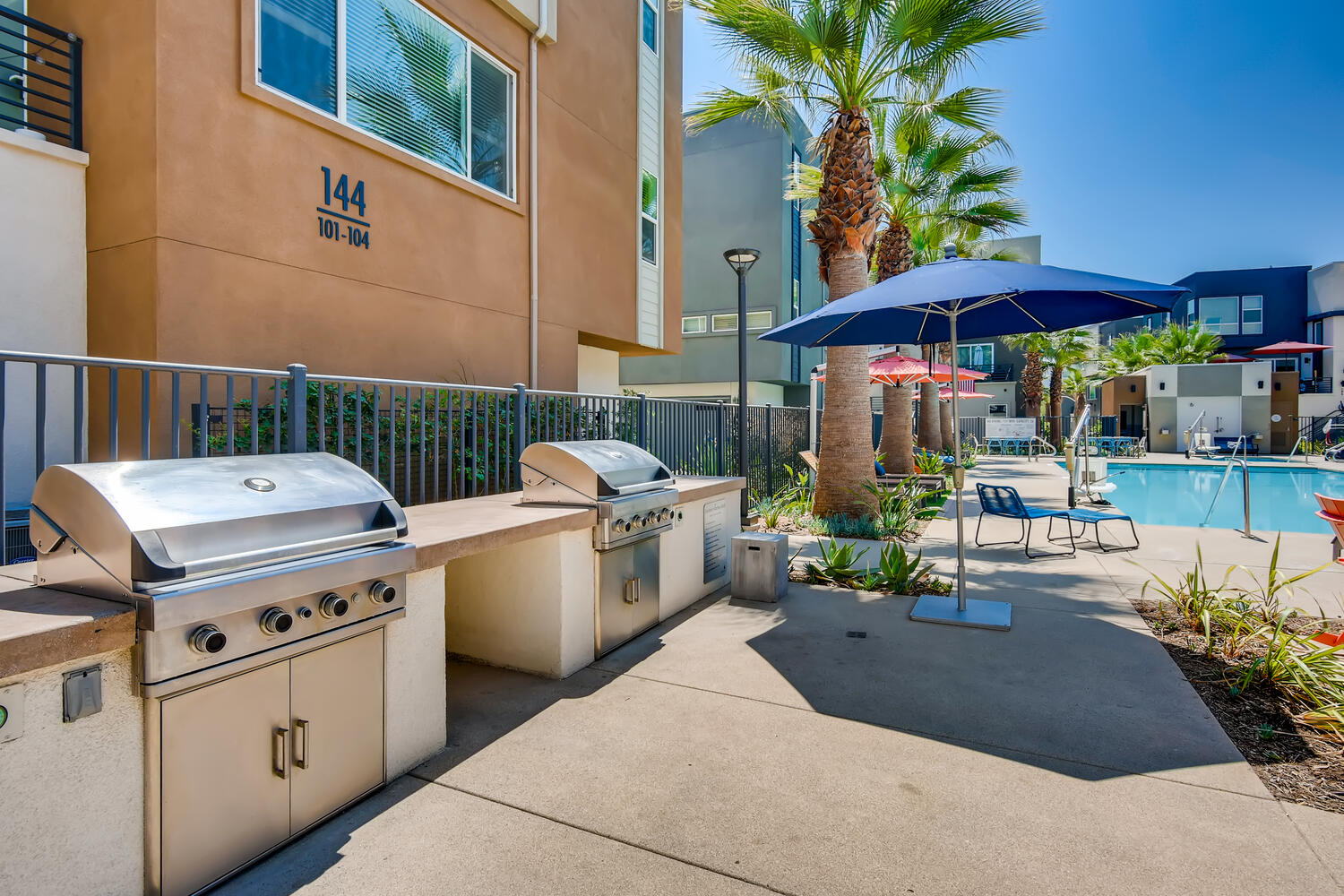 Outdoor pool area in a residential complex with stainless steel grills, a blue umbrella, and a black metal fence.