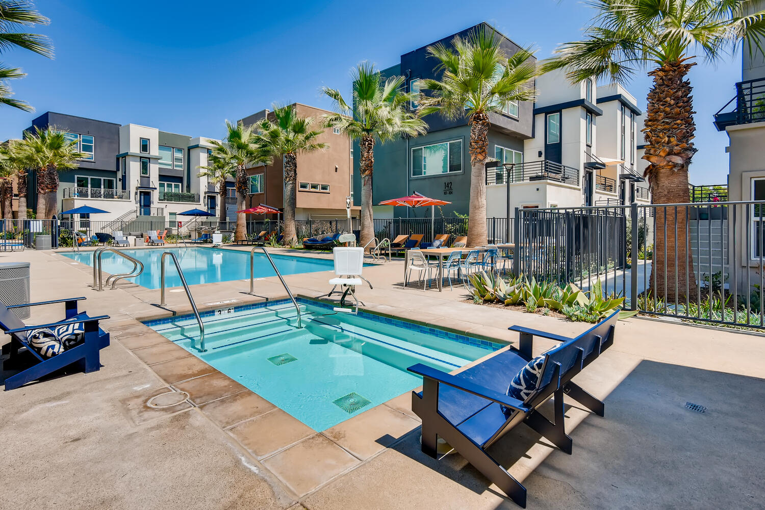 swimming pools with lounge chairs, umbrellas, palm trees, and a metal fence.