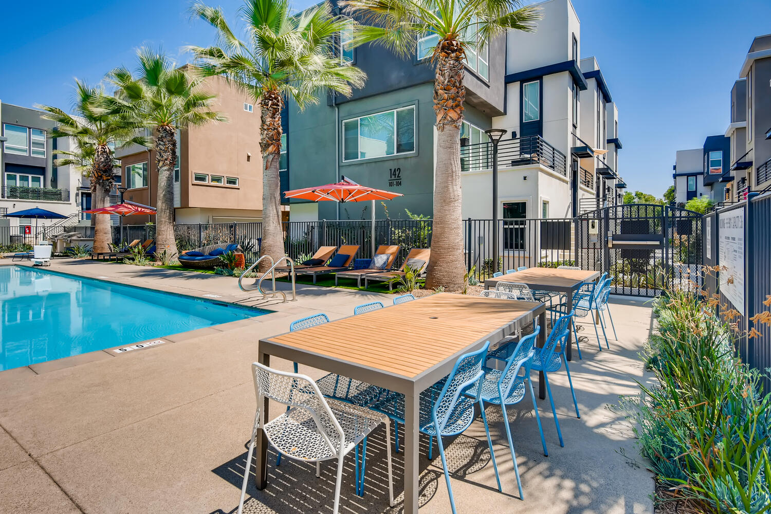 pool with lounge chairs, umbrellas, and a wooden table with blue chairs.